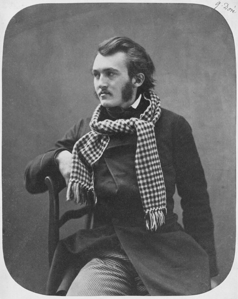 Black and white photograph of the young Gustave Doré, seated sideways on a chair with his arm over its back, wearing a dark coat and a checked scarf, looking to the left.