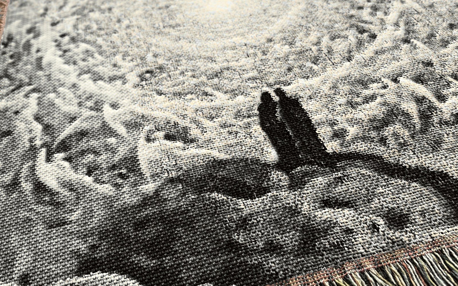 Close-up of the woven Paradiso blanket in gray and cream: two small dark figures on a rock beneath rings of angels and a soft glow, above a colorful fringe.