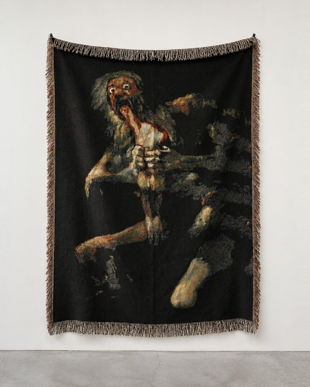 Saturn woven blanket hung on a white gallery wall: the god gripping a pale figure against a black ground, fringed edges.