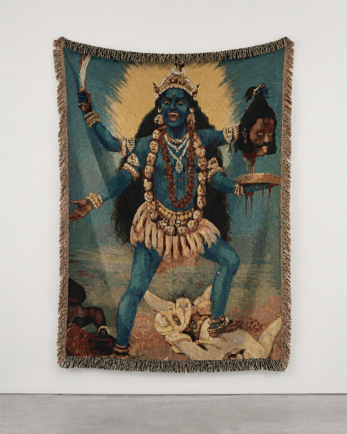 Kali wall hanging on a white gallery wall: the four-armed blue goddess before a golden halo, fringed edges.