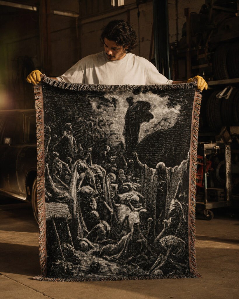 A man in a white T-shirt and yellow work gloves holds up the woven Valley of Dry Bones blanket in a workshop, showing its full length; the black and grey image with fringed edges reaches down to the concrete floor.