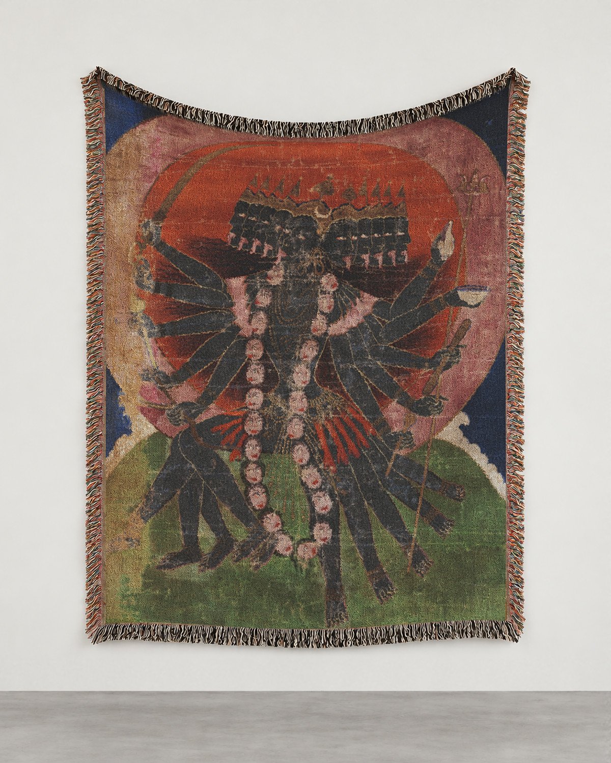 Goddess Mahakali wall hanging on a white gallery wall: the blue-black goddess with ten heads under one crown, on a green hill before a great red disc, fringed edges.